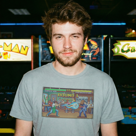 Man standing in a retro arcade wearing a coffee barista inspired arcade game t-shirt in grey