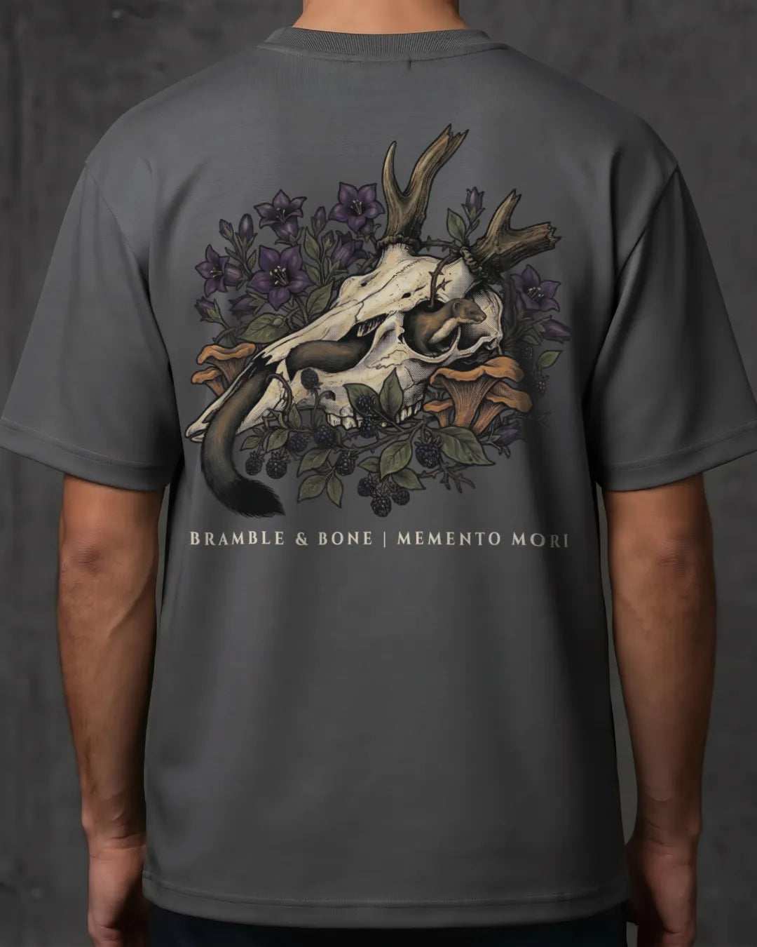 A man wearing a dark gray t-shirt, viewed from the back. The shirt features a large graphic print of a deer skull with antlers, surrounded by purple flowers, green leaves, and brown branches, with the text 'BRAMBLE & BONE | MEMENTO MORI' printed below.