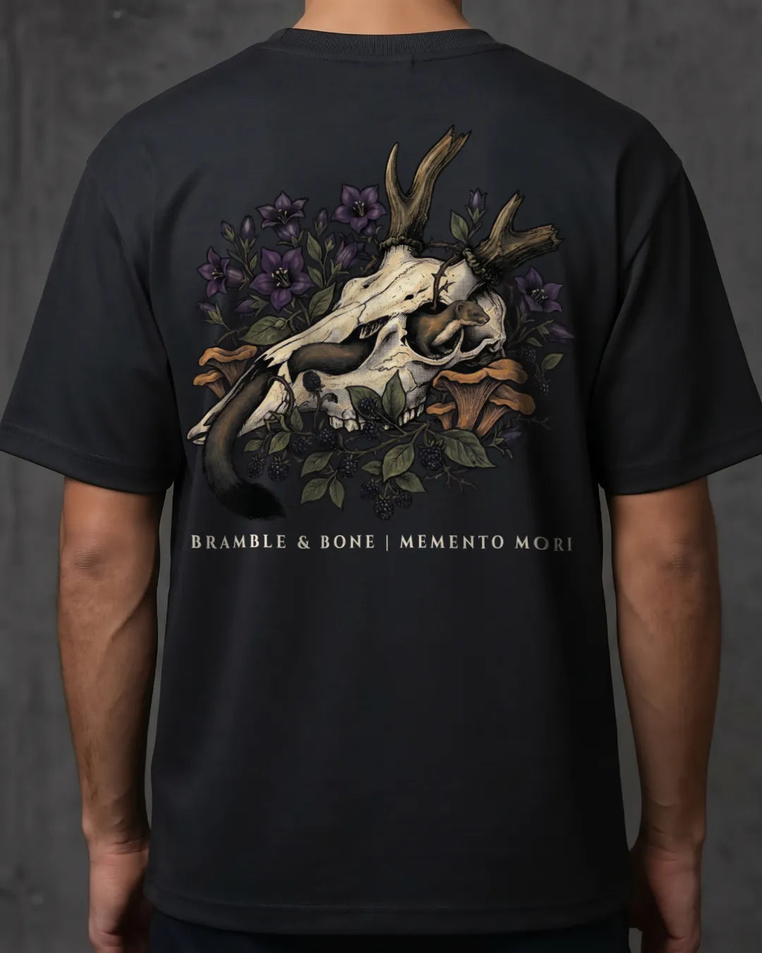 A person wearing a black short-sleeve T-shirt shown from the back, featuring a large graphic print of a deer skull with antlers, purple flowers, green leaves, and a stoat, with the text 'BRANBLE & BONE | MEMENTO MORI' printed below.