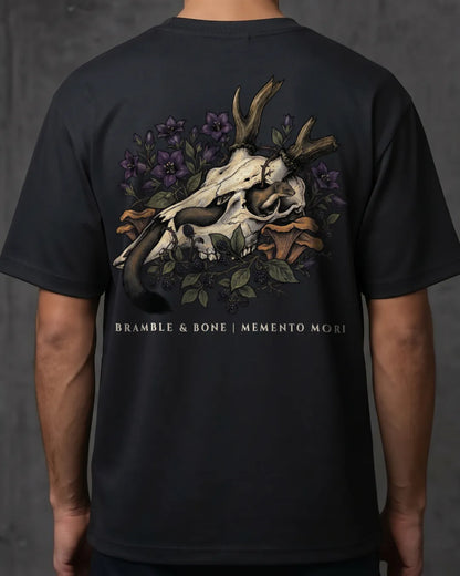 A person wearing a black short-sleeve T-shirt shown from the back, featuring a large graphic print of a deer skull with antlers, purple flowers, green leaves, and a stoat, with the text 'BRANBLE & BONE | MEMENTO MORI' printed below.