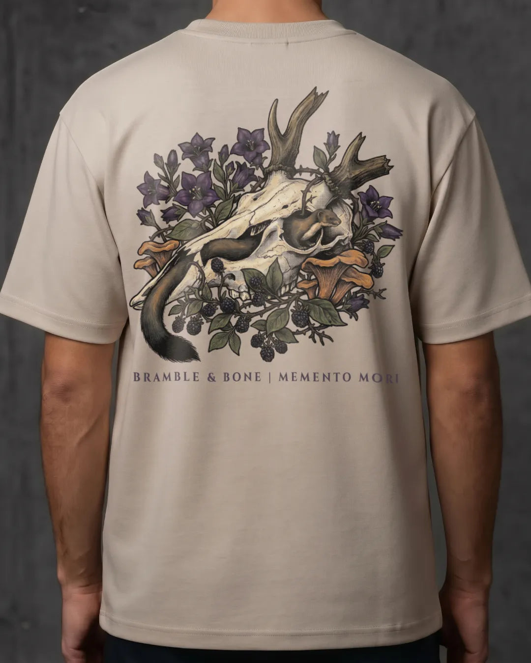 A person wearing a light beige short-sleeve T-shirt, shown from the back. The shirt features a large graphic print of a deer skull surrounded by purple flowers, green leaves, and orange mushrooms, with the text 'BRANBLE & BONE | MEMENTO MORI' printed below.