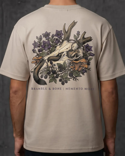 A person wearing a light beige short-sleeve T-shirt, shown from the back. The shirt features a large graphic print of a deer skull surrounded by purple flowers, green leaves, and orange mushrooms, with the text 'BRANBLE & BONE | MEMENTO MORI' printed below.