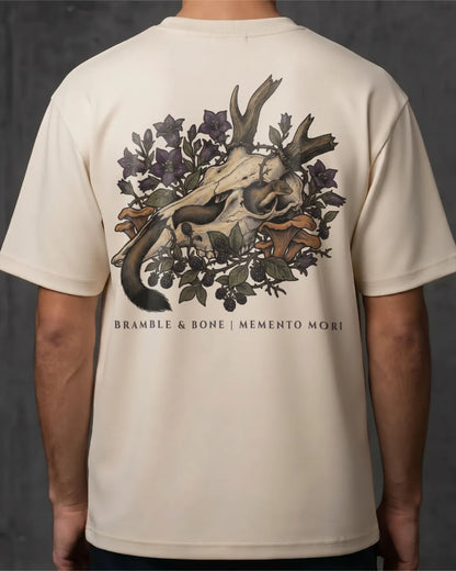 A person wearing a cream-colored t-shirt, showing the back of the garment with a large graphic print of a deer skull, antlers, leaves, and flowers, with the text 'BRANBLE & BONE | MEMENTO MORI' printed below.