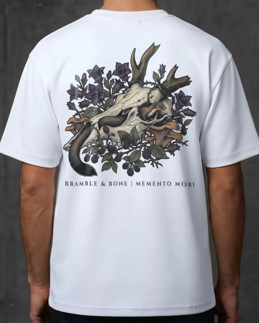 A white short-sleeve t-shirt shown from the back on a model, featuring a large graphic print of a skull with antlers surrounded by dark purple flowers and green leaves, with the text 'BRAMBLE & BONE | MEMENTO MORI' printed below.