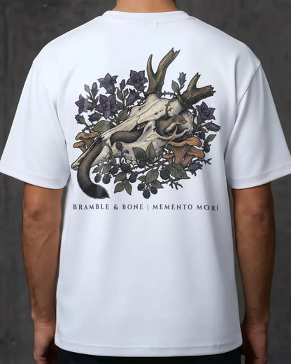 A white short-sleeve t-shirt shown from the back on a model, featuring a large graphic print of a skull with antlers surrounded by dark purple flowers and green leaves, with the text 'BRAMBLE & BONE | MEMENTO MORI' printed below.