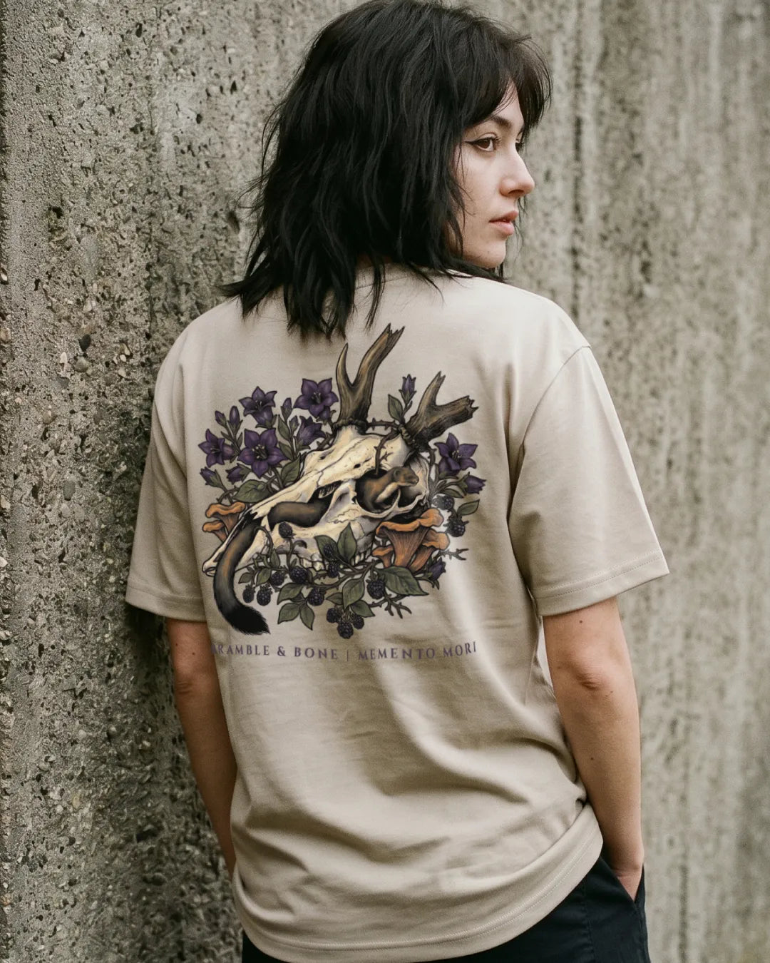 A woman with dark hair is shown from behind, wearing a beige t-shirt with a large graphic print on the back featuring a deer skull, antlers, purple flowers, and a snake. The text 'Bramble & Bone' is printed below the graphic.