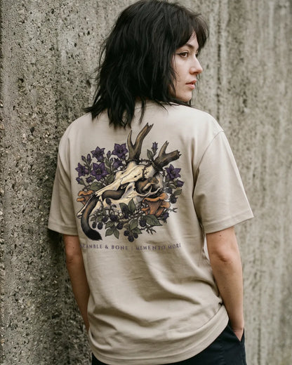A woman with dark hair is shown from behind, wearing a beige t-shirt with a large graphic print on the back featuring a deer skull, antlers, purple flowers, and a snake. The text 'Bramble & Bone' is printed below the graphic.