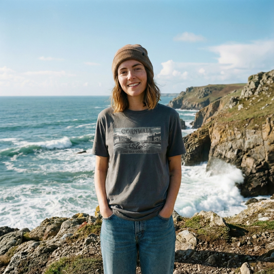 Model on a Cornish cliffside wearing a grey 'The Old Gods Wait' t-shirt. The graphic features a vintage-style travel poster of a Cornish harbour with a subtle cosmic horror influence.