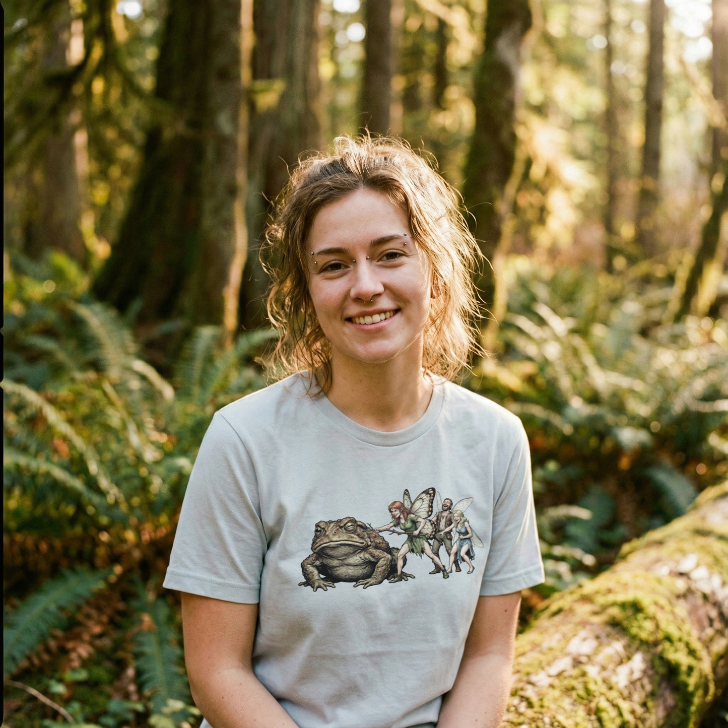 Graphic tee featuring a fairy poking a giant grumpy toad with a stick while other fairies laugh, organic cotton fantasy t-shirt.