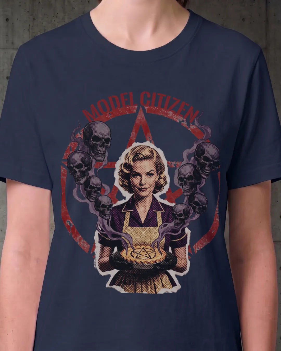 A navy blue short-sleeve t-shirt with a vintage-style graphic print of a woman holding a pie, surrounded by skulls and purple smoke, with the text 'MODEL CITIZEN' arched above. The shirt is shown on a model against a concrete wall background.