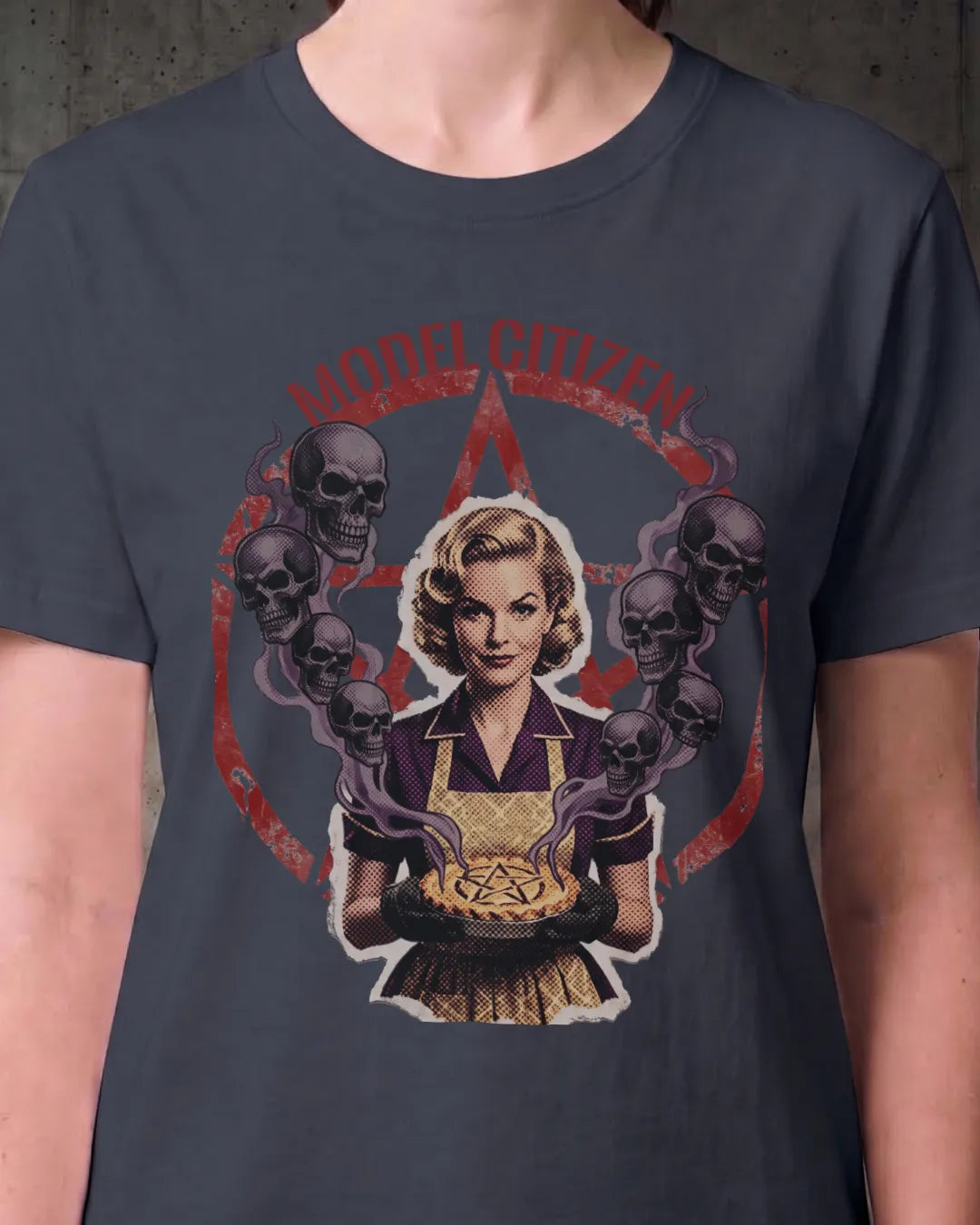 A person wearing a dark charcoal gray t-shirt with a graphic print. The graphic features a retro-style illustration of a blonde woman in a kitchen holding a pie, surrounded by skulls and purple smoke, with the text 'MORE CIVILIAN' arched above her against a red pentagram.