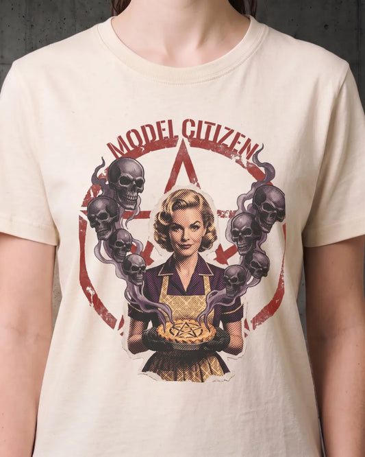 A person wearing a cream-colored graphic t-shirt. The shirt features a retro-style pin-up illustration of a woman holding a pie, surrounded by skulls and a red pentagram, with the text 'MODEL CITIZEN' arched above her.
