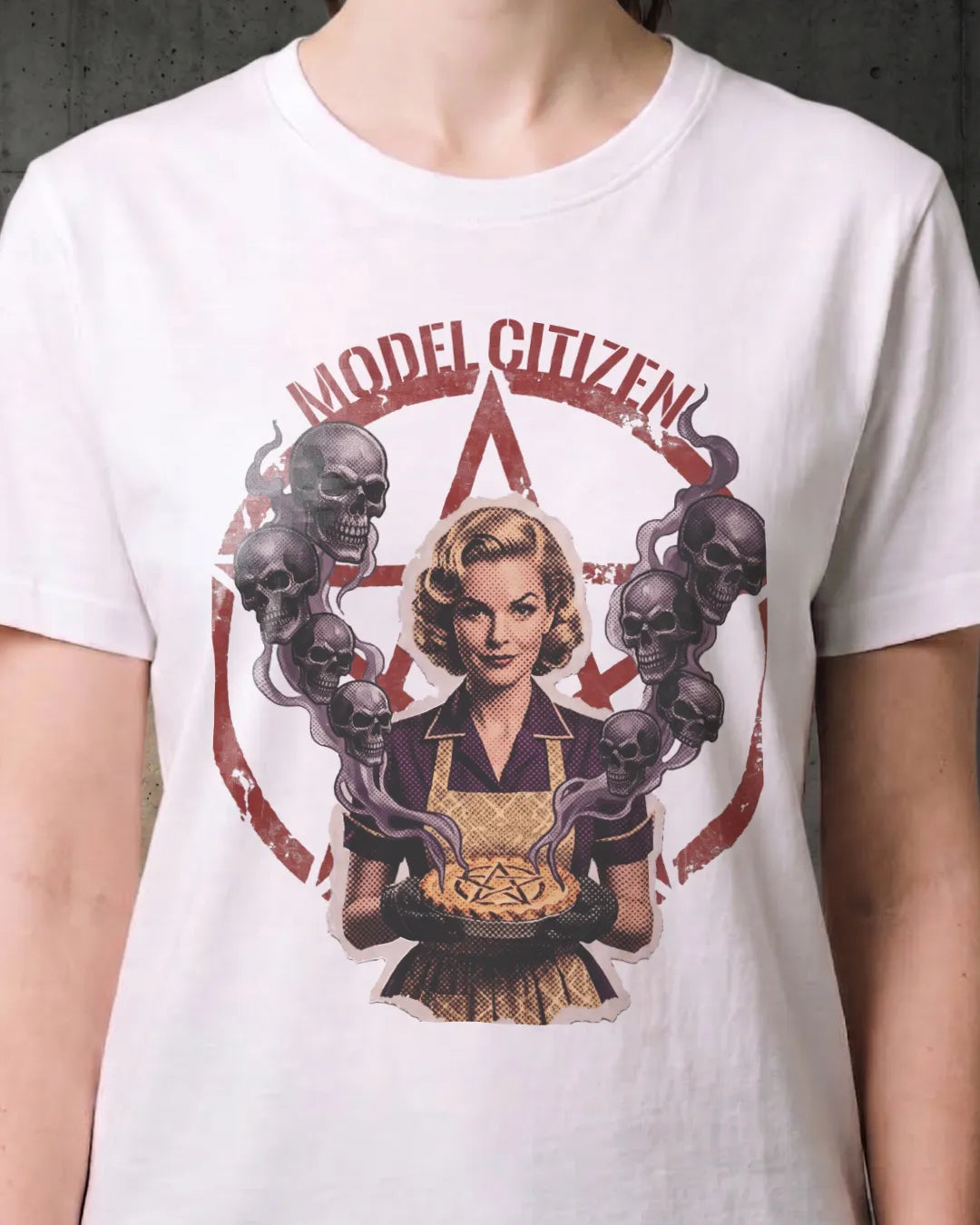 A white short-sleeve graphic t-shirt worn by a model, featuring a retro-style illustration of a blonde woman in a kitchen apron holding a pie, surrounded by skulls and a red pentagram, with the text 'MODEL CITIZEN' arched above.