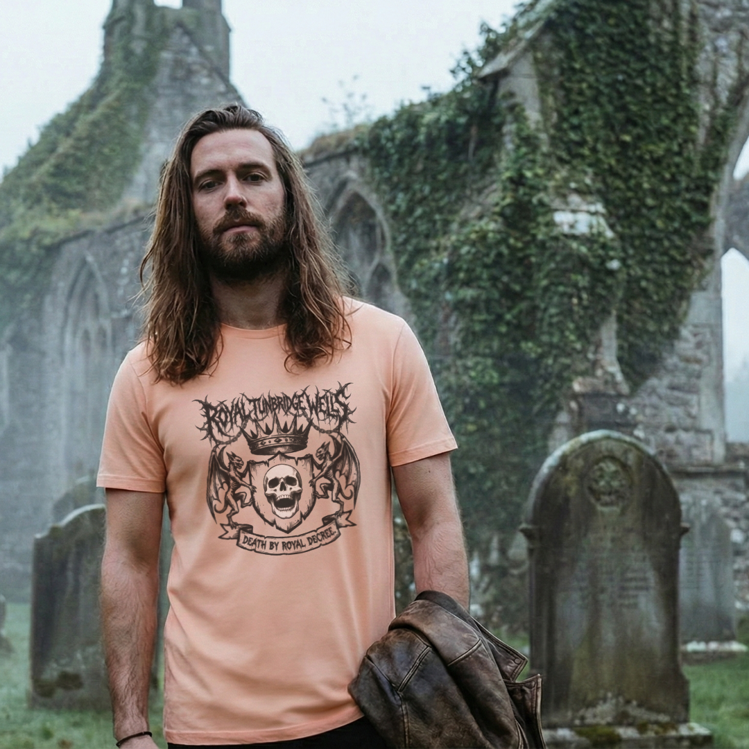 Model in a historic graveyard setting wearing a peach-colored 'Royal Tunbridge Wells' t-shirt. The graphic features a brutal black metal-style illustration with demonic gargoyles and a screaming skull.
